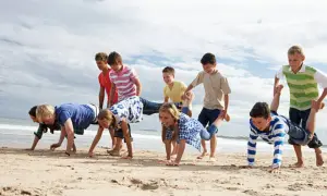 Niños compiten en juegos en la playa sin material, haciendo carreras de carretilla humana y otros desafíos divertidos.