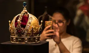 Złota korona, ozdobiona perłami i rubinami, przypomina wielki mogoł diament. Kobieta w okularach robi jej zdjęcie telefonem.