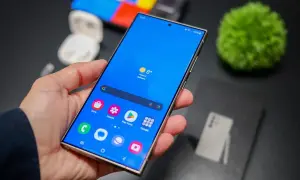 Mano sujetando un smartphone con pantalla azul vibrante, mostrando iconos de aplicaciones y el tiempo. La interfaz, **que es One UI**, se ve fluida y organizada.
