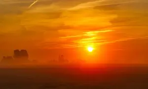 Een gloedvolle zonsopgang boven een mistig veld. De zon, een heldere bol, kondigt het wakker worden van de dag aan, met oranje en gele tinten die de hemel vullen.