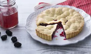 Une délicieuse tarte aux mûres, avec une part coupée, sur une assiette ornée. Des mûres fraîches et un verre de jus rouge complètent la scène.