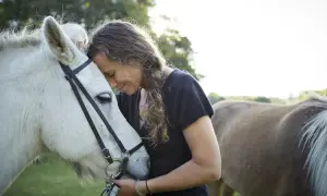 Kobieta przytula się do białego konia, czując jego ciepło. W tle widać drugiego konia. To piękny widok więzi między człowiekiem a zwierzęciem, gdzie szory dla konia są tylko elementem otoczenia.