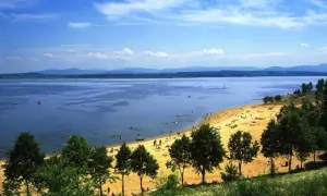 Słoneczna plaża nad Jeziorem Nyskim, pełna ludzi odpoczywających i kąpiących się. W oddali góry, a na wodzie żaglówki i łodzie. To idealne miejsce na wakacyjne atrakcje.