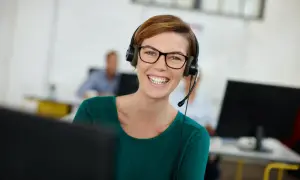 Uśmiechnięta kobieta w okularach i zestawie słuchawkowym, korzystająca z technologii VoIP w biurze.