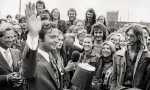 Kung Carl XVI Gustaf vinkar glatt till en folkmassa. En ung kvinna håller fram en mikrofon, kanske för att intervjua en av kungens barn eller någon annan i den glada gruppen.