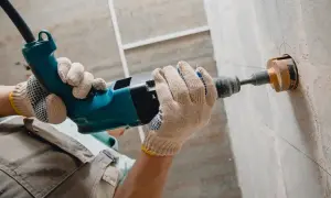 Ręce w rękawicach wiercą otwór w ścianie wiertarką udarową. To kluczowy etap prac, zgodny z przepisami dotyczącymi wkuwania rur w ścianę.