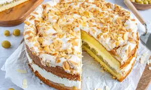 Ein köstlicher Stachelbeer Baiser Kuchen mit Biskuitböden, Sahne und Mandelsplittern. Ein Stück ist bereits angeschnitten.