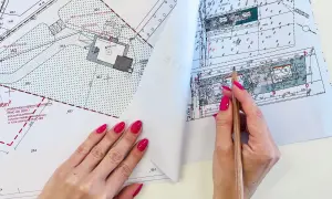 Dłoń z różowym lakierem na paznokciach wskazuje ołówkiem na mapę projektową z zaznaczonym budynkiem i wymiarami.