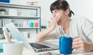 Kobieta z kubkiem kawy ziewa przy laptopie, czując abulię i brak motywacji do pracy.