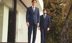 Zwei Männer in eleganten Anzügen stehen auf einer Holzterrasse. Der vordere Mann zeigt, wie man einen Anzug richtig trägt, mit perfekter Passform und Stil.