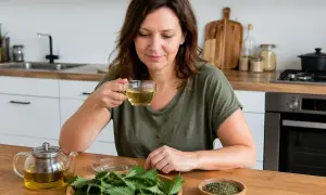 Frau trinkt Brennnesseltee in der Küche. Sie fragt sich, ob Brennnesseltee beim Abnehmen hilft. Frische Brennnesseln und getrocknete Kräuter liegen auf dem Tisch.