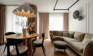 Przytulny salon z beżową kanapą i okrągłym stołem. Nad kanapą w salonie wiszą dekoracyjne lampy, a obok stoi zielona pufa.