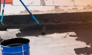 Osoba wykonuje hydroizolację piwnicy od wewnątrz, malując ściany czarną masą bitumiczną za pomocą wałka.