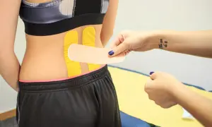 Naklejanie tejpy na plecy w odcinku lędźwiowym. Dłoń z tatuażem "You can do it" przygotowuje plaster.