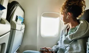 Vrouw met oortjes kijkt uit het vliegtuigraam, hopend op tips wat te doen tegen vliegangst. Ze houdt haar telefoon vast.