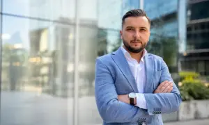 Mężczyzna w jasnoniebieskiej marynarce i białej koszuli, z rękami skrzyżowanymi na piersi, na tle nowoczesnego budynku.