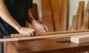 Manos midiendo una tabla de madera en un taller. Se aprecian otras tablas en el fondo.