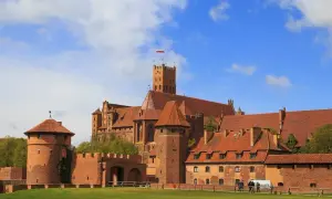 Malbork, jeden z największych zamków w Polsce, z charakterystyczną wieżą i czerwonymi dachami, na tle błękitnego nieba z chmurami.