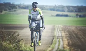 Kolarz w stroju sportowym jedzie rowerem po szutrowej drodze wśród pól.