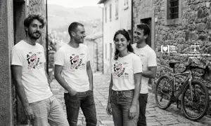 Quattro amici sorridono in un vicolo di pietra, indossando magliette con un disegno di bici e la scritta "A tutta virra". Un'espressione del vocabolario dialetto marchigiano che evoca passione.