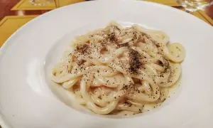 Piatto di pici cacio e pepe, pasta fresca condita con pecorino romano e pepe nero macinato fresco, servita su un piatto bianco.
