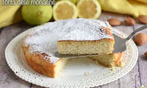 Torta delle monache soffice e profumata, cosparsa di zucchero a velo, con limoni e mandorle sullo sfondo.