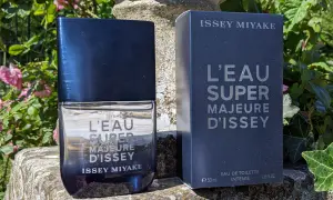 Flakon i pudełko perfum Issey Miyake L'Eau Super Majeure d'Issey na kamiennym postumencie, otoczone zielenią i różowymi kwiatami.