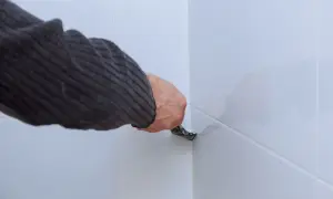 Ręka w swetrze usuwa silikon z białych paneli podłogowych za pomocą narzędzia.