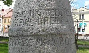 Kamienny słup milowy konin z wyrytymi łacińskimi napisami, na tle budynków i zieleni.