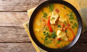 Bol de blanquette de poisson crémeuse avec des morceaux de poisson, des poivrons rouges et du persil, servi sur une serviette jaune sur une table en bois.