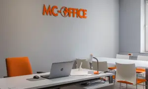 Pomarańczowe logo "MC-OFFICE" na szarej ścianie, biurko z laptopem i krzesło.
