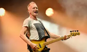 Sting, lider zespołu The Police, gra na gitarze basowej na scenie, oświetlony ciepłym światłem.