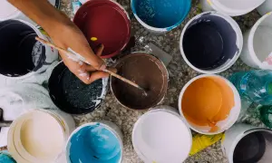Ręka z pędzlem miesza farby w wiaderkach. Widać różne kolory, co może sugerować wady farby ceramicznej, np. nierównomierne krycie.