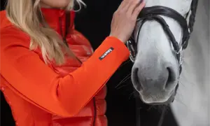 Pomarańczowa kurtka na konie z logo Eskadron. Kobieta głaszcze białego konia.