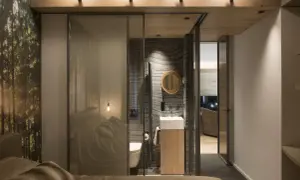 Bagno in camera di 2 mq con pareti in pietra, lavabo sospeso e specchio rotondo. La zona notte è separata da una parete in vetro con stampa foresta.