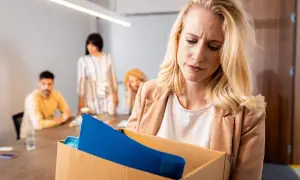Femme blonde triste portant une boîte de ses affaires, se demandant si l'on peut licencier sans motif. Des collègues flous en arrière-plan.