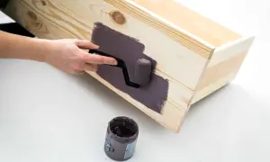 En hand använder en liten roller för att måla en träkista med mörklila färg, vilket ger en jämn yta utan synliga penseldrag.