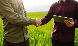 Dwóch mężczyzn w polu ściska sobie dłonie, jeden z nich trzyma tablet. Rozmowa o tym, jak sprawdzić, czy działka jest uzbrojona.