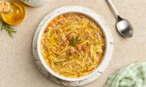 Un bol de pasta e fagioli, une soupe réconfortante avec des pâtes fines et des haricots, garnie de romarin. Une cuillère et une petite bouteille d'huile d'olive sont à proximité.