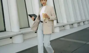 Eine Frau im smart casual Look mit beigem Blazer, Jeans und Kaffee in der Hand, geht mit einem Tablet.