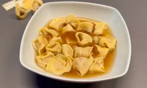 Un piatto fumante di tortellini in brodo, un classico confortante. Sullo sfondo, alcuni tortellini crudi e sfoglia.