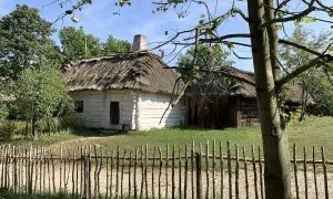 Drewniane chaty kryte strzechą w skansenie, przed nimi płotek z gałęzi.
