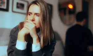 Kobieta zamyślona, z rękami przy twarzy, w tle niewyraźna postać. Może to symbolizować dystans emocjonalny w pracy, gdy czujemy się odizolowani.