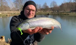 Glücklicher Angler hält frisch gefangene Forelle am Forellenteich. Der Fisch hat einen grellgrünen Köder im Maul.