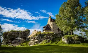 Kamienny zamek na skale, z polską flagą, w otoczeniu zieleni i skalnych formacji. W oddali widać łuk skalny, przez który przechodzą turyści. To malownicze mirów bobolice.