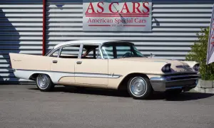 En krämvit och ljusbeige De Soto från 1950-talet står parkerad framför en byggnad med skylten "AS CARS".