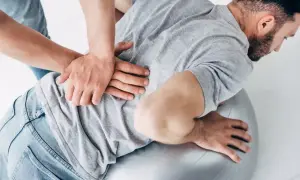 Terapeuta wykonuje masaż pleców mężczyzny na piłce rehabilitacyjnej. Czy osteopata jest lekarzem? To pytanie nurtuje wielu pacjentów.