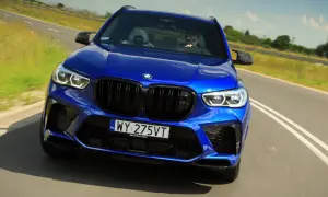 Niebieski BMW X5 M Competition pędzi po drodze, kierowca w okularach przeciwsłonecznych za kierownicą.