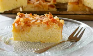 Ein Stück saftiger **Mandelkuchen vom Blech** mit knusprigen Mandelblättchen und Zucker bestreut, auf einem Glas-Teller mit einer Gabel.