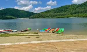 Płytkie jezioro dla dzieci z kolorowymi rowerami wodnymi i kajakami na piaszczystej plaży, otoczone zielonymi wzgórzami.